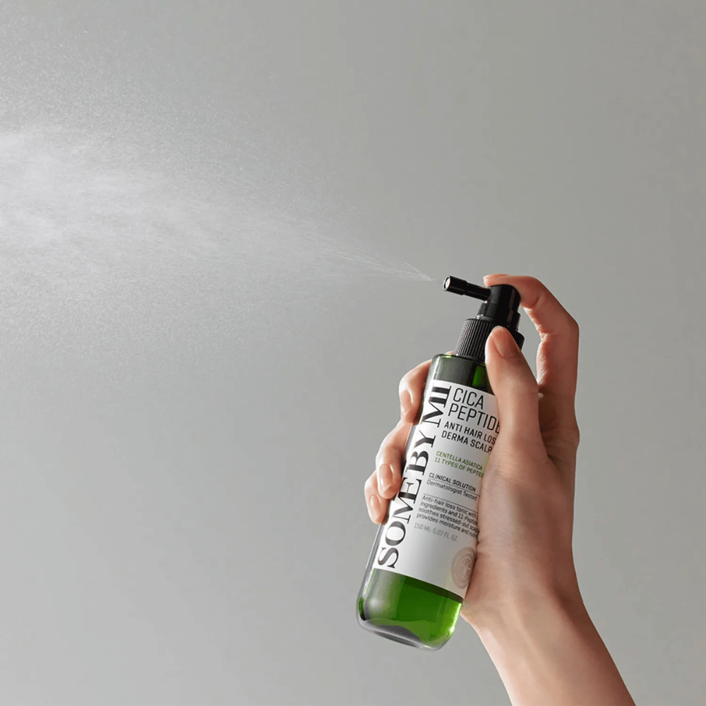 Hånd som sprayer SOME BY MI Cica Peptide Anti Hair Loss Derma Scalp Tonic fra en grønn flaske med svart munnstykke og hvit etikett mot grå bakgrunn.