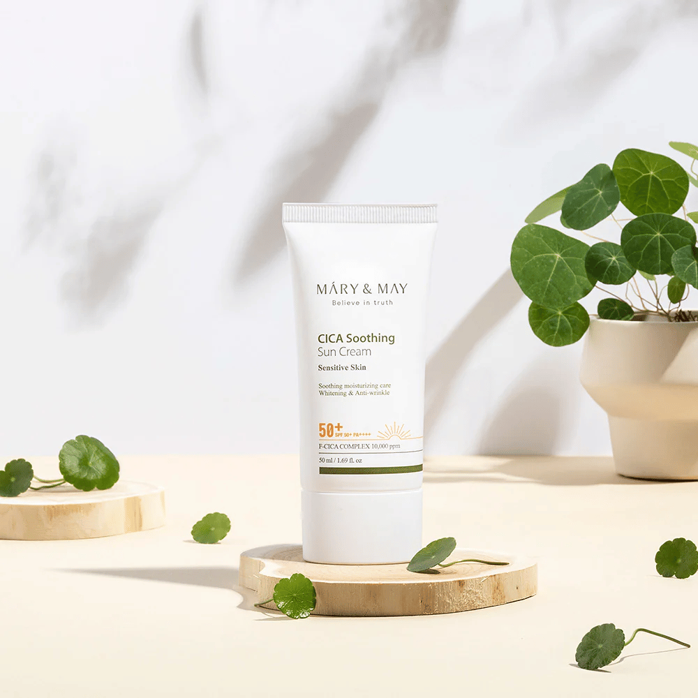 Mary&May CICA Soothing Sun Cream plassert på et trepodium med grønne centella-blader rundt omkring. Bildet understreker de hudberoligende ingrediensene og den naturlige inspirasjonen bak produktet.