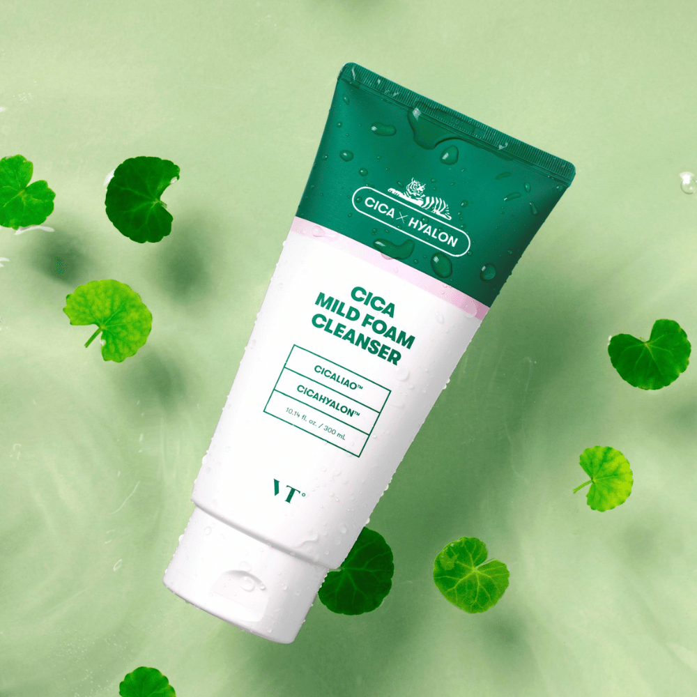 VT Cica Mild Foam Cleanser liggende mot et lysegrønt underlag med vanndråper på emballasjen og omgitt av grønne centella asiatica-blader, noe som fremhever produktets beroligende egenskaper.
