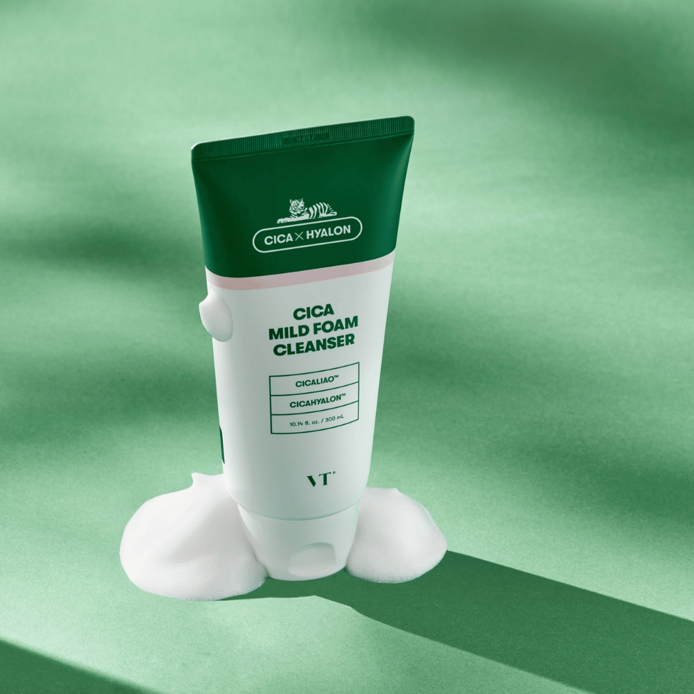 Tube med VT Cica Mild Foam Cleanser stående på et grønt underlag med skum rundt basen. Produktet har grønn og hvit design og markedsføres med CICALIAO™ og CICAHYALON™.
