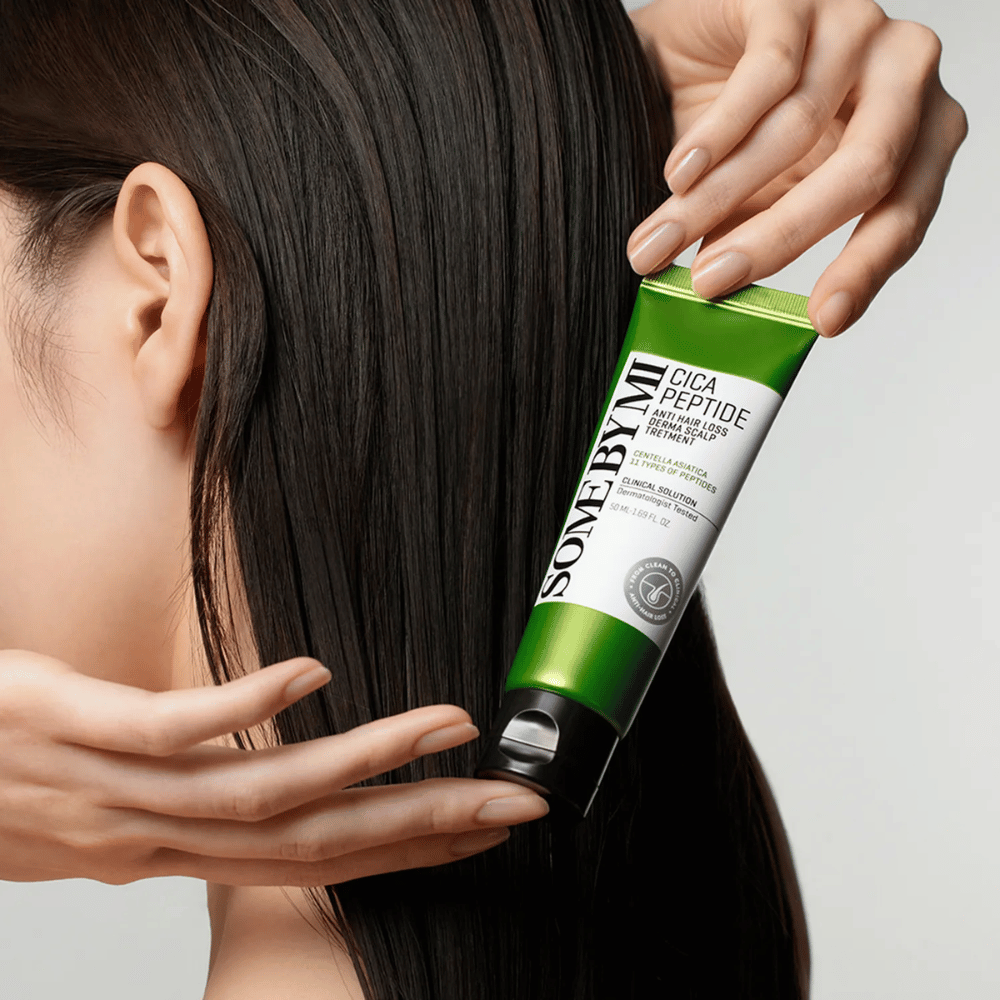 Kvinne påfører SOME BY MI Cica Peptide Anti Hair Loss Derma Scalp Treatment i langt, mørkt hår med en grønn og hvit tube.