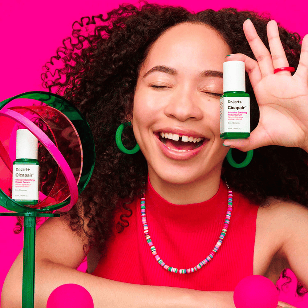 En kvinne smiler bredt mens hun holder Dr.Jart+ Cicapair Intensive Soothing Repair Serum foran ansiktet sitt. Hun bærer grønne øredobber og et rødt halskjede. Bakgrunnen er lys rosa og dekorert med grønne og rosa sirkler.