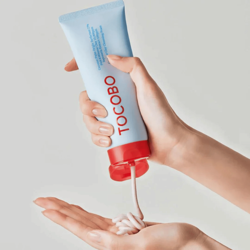 En person holder i en tube av TOCOBO Coconut Clay Cleansing Foam. Tuben er lyseblå med en rød kork og teksten "TOCOBO" på forsiden. Produktet trykkes ut av tuben og formes som en sjampo i hånden. Bakgrunnen er nøytral og gir fokus på produktet.