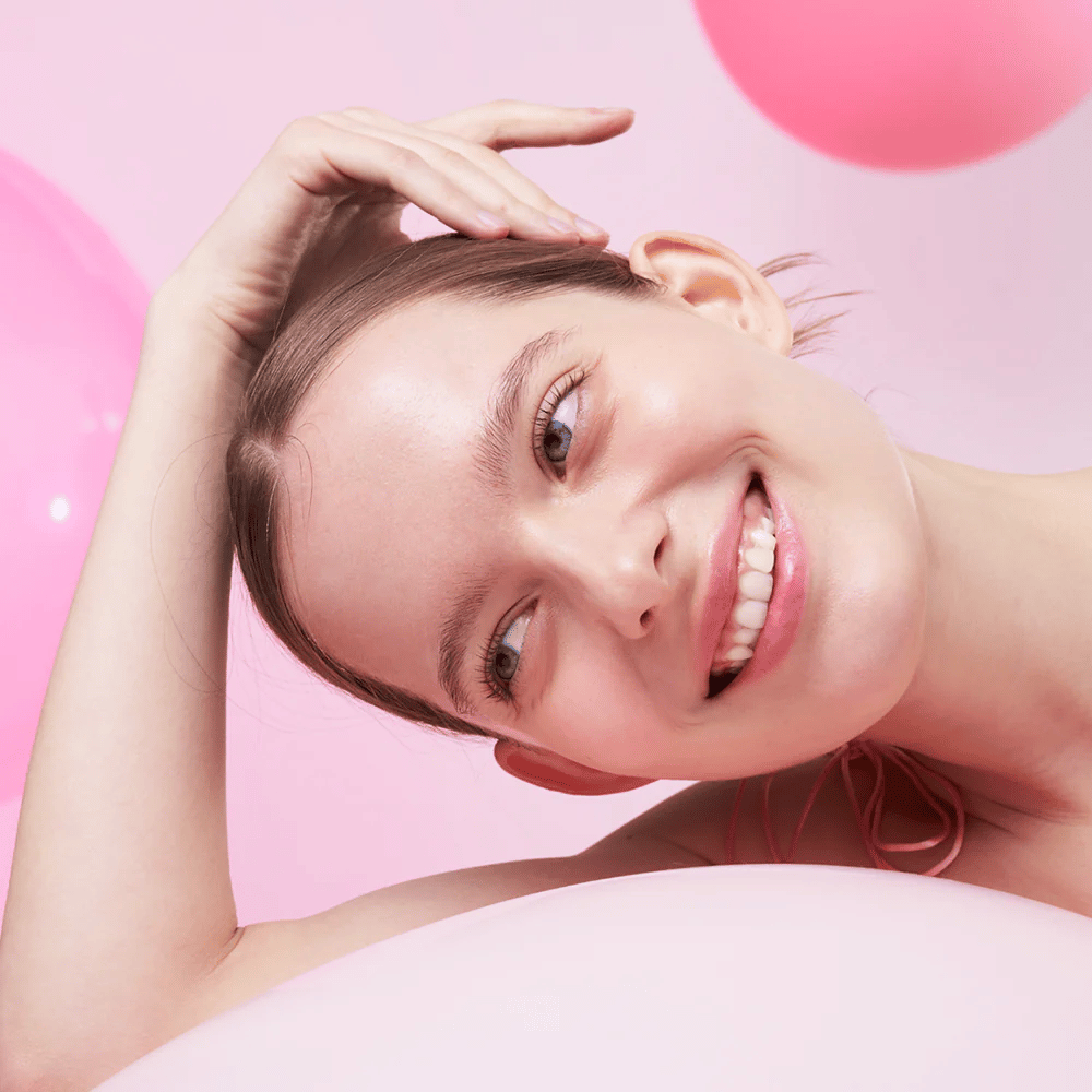 En kvinne smiler og hviler hodet på hånden, omgitt av rosa luftballonger. Huden hennes ser strålende og naturlig frisk ut.