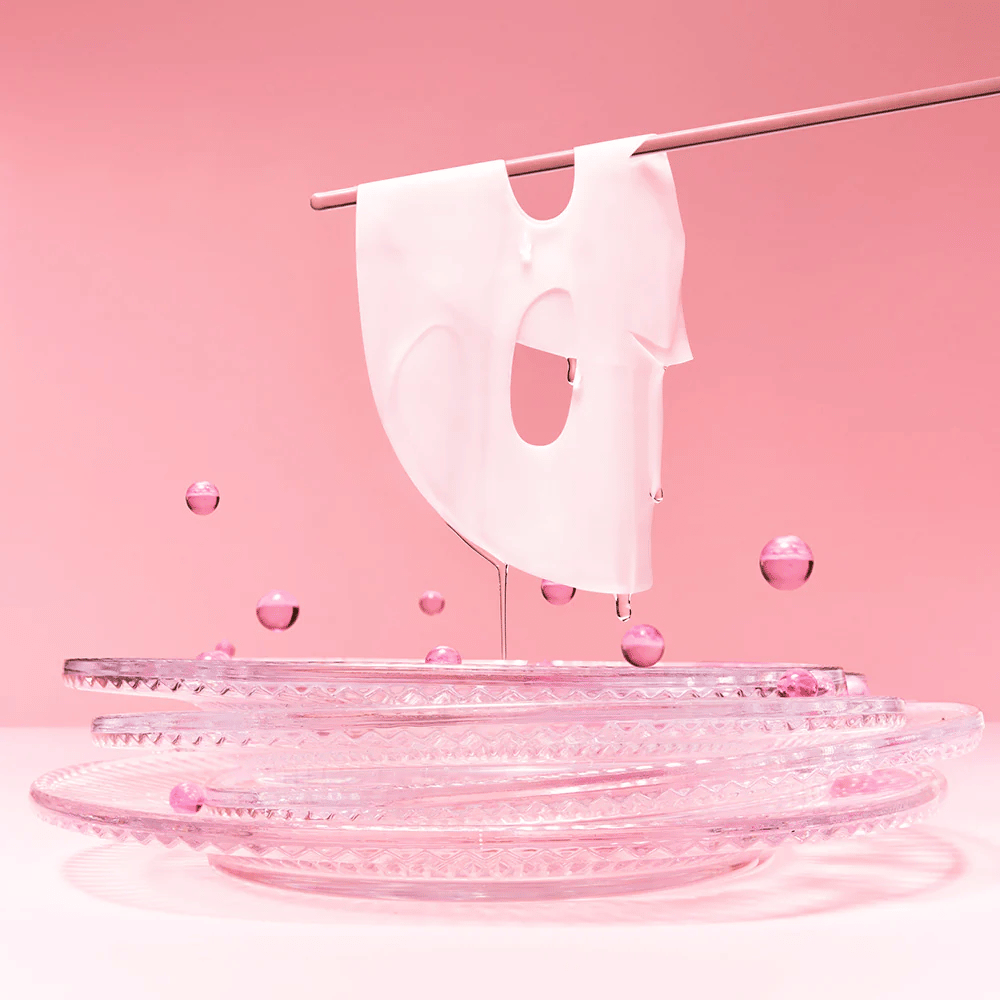 En hydrogelmaske henger på en stang, dryppende med serum. Bakgrunnen er rosa og speilet på bunnen reflekterer de rosa boblene som omgir masken.