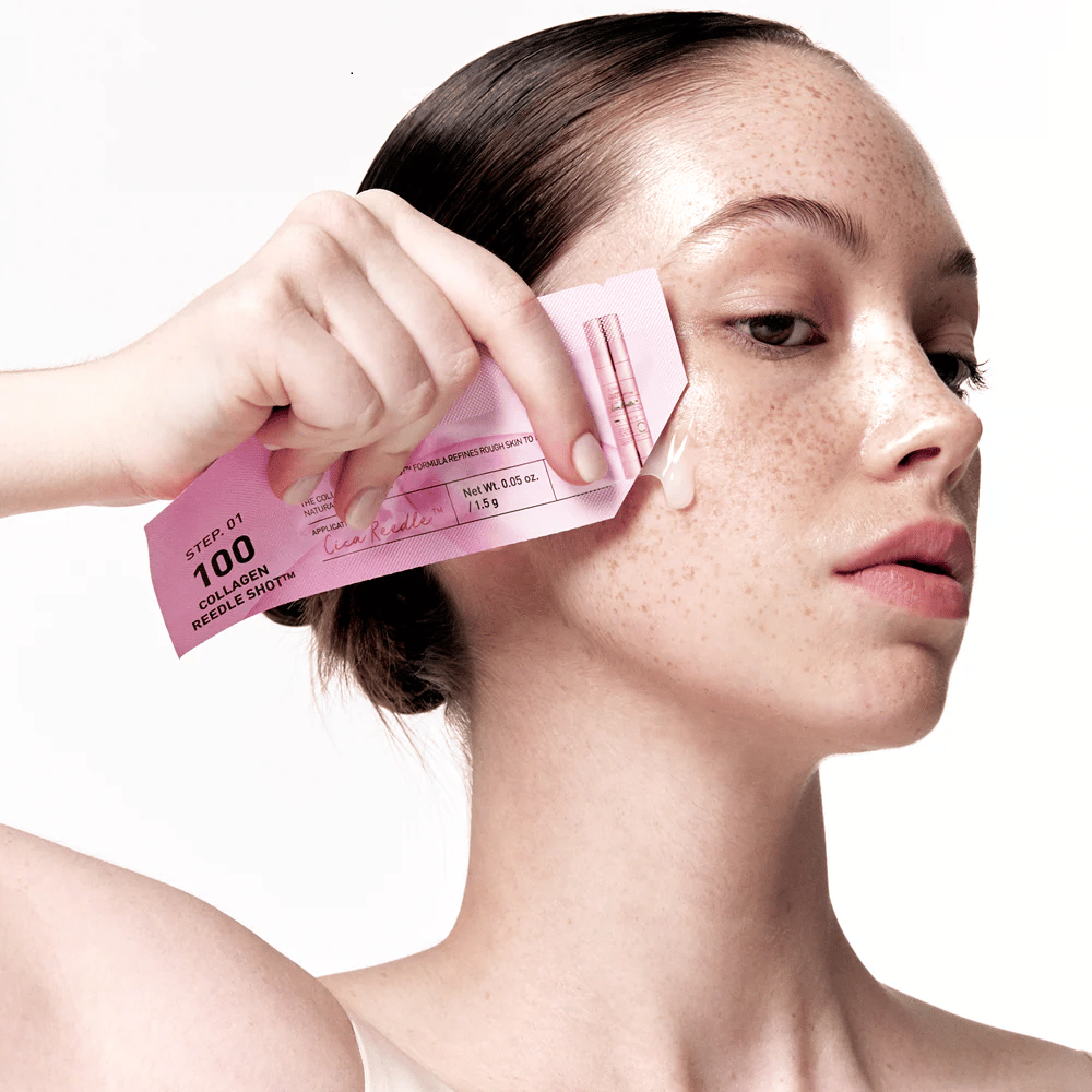 Kvinne påfører trinn 1 av Collagen Reedle Shot 100 med en rosa engangspakning på kinnet. En klar gel vises på huden, noe som markerer det første trinnet i den totrinns hudpleiebehandlingen.