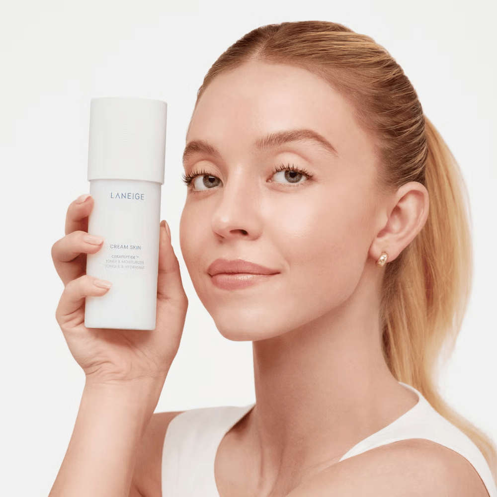 En person holder opp Laneige Cream Skin Cerapeptide Refiner i en hvit flaske. Personen smiler og viser produktet, som markedsføres som en fuktighetsgivende toner og hudpleieprodukt.