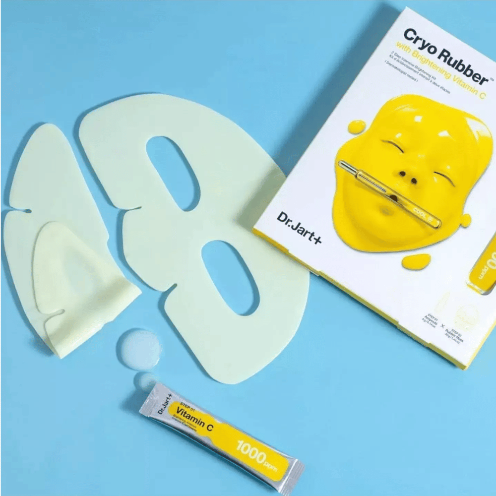 Bilde av Dr.Jart+'s 'Cryo Rubber™ med Brightening Vitamin C' skjønnhetsprodukt. På en blå bakgrunn sees en hvit gummiansiktsmaske, en liten tube merket 'Vitamin C 1000ppm', og en eske med produktets navn og bilde av den gule masken.