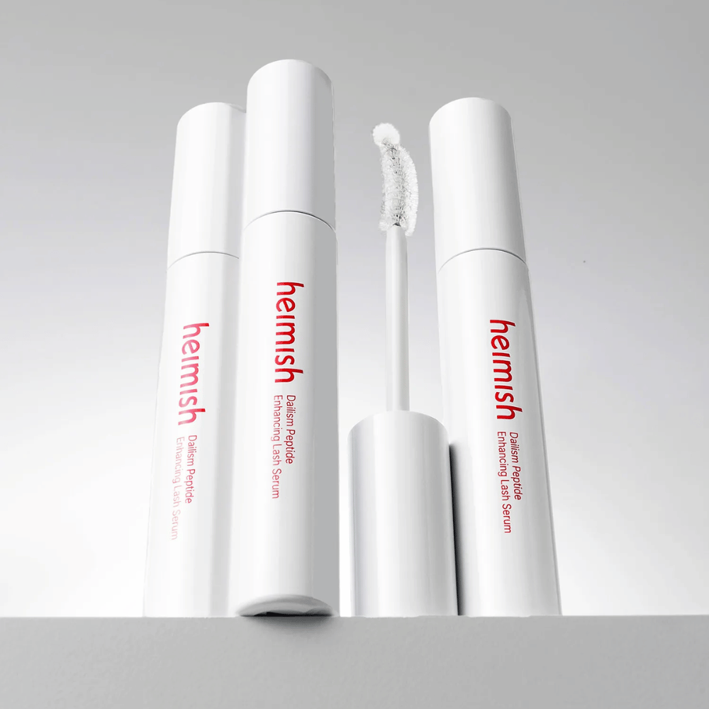 Flere pakninger av Heimish Dailism Peptide Enhancing Lash Serum vises sammen med den buede applikatorbørsten. Bildet fremhever produktets elegante design og presisjonsbørste for enkel påføring.