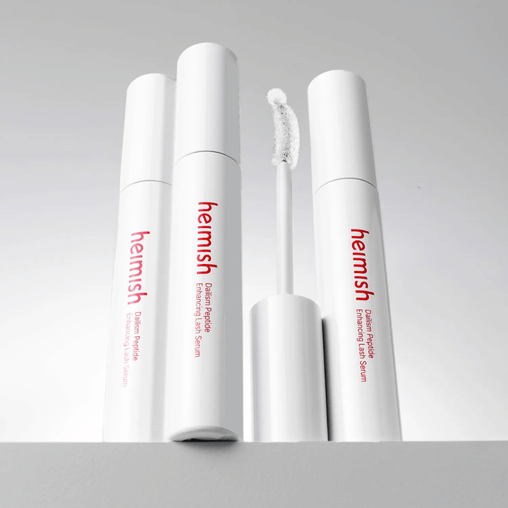 Flere pakninger av Heimish Dailism Peptide Enhancing Lash Serum vises sammen med den buede applikatorbørsten. Bildet fremhever produktets elegante design og presisjonsbørste for enkel påføring.