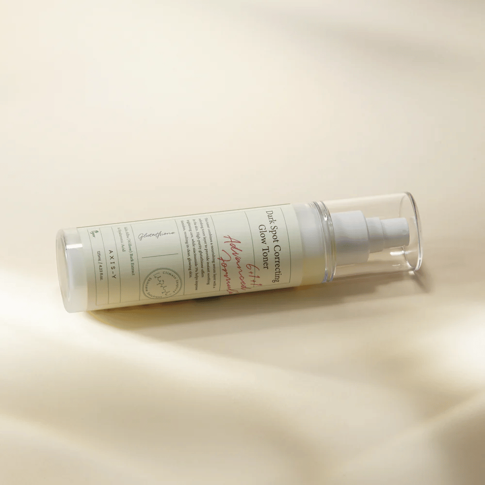 En liggende flaske av "AXIS-Y Dark Spot Correcting Glow Toner" plassert på en lys overflate. Flasken er gjennomsiktig med en hvit etikett og har en hvit spraypumpe. Etiketten viser informasjon om produktets lysnende og fuktighetsgivende egenskaper samt inneholder nøkkelingredienser som Glutathione og Niacinamide.