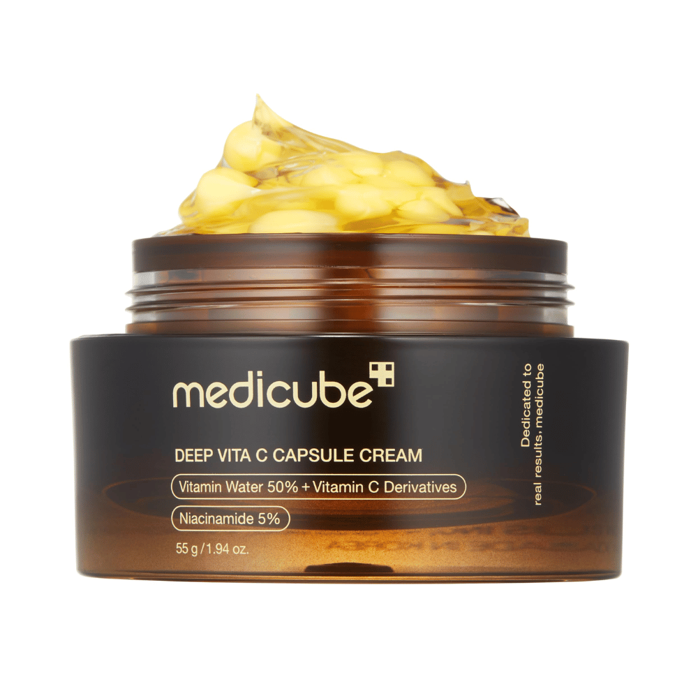 Et nærbilde av "Medicube Deep Vita C Capsule Cream," som viser kremens konsistens og kapslene av vitamin C. Emballasjen er mørkebrun med gulltekst og beskriver innholdet av vitaminvann (50 %) og niacinamid (5 %), som gir en visuell presentasjon av produktets fuktighetsgivende og hudforbedrende egenskaper.