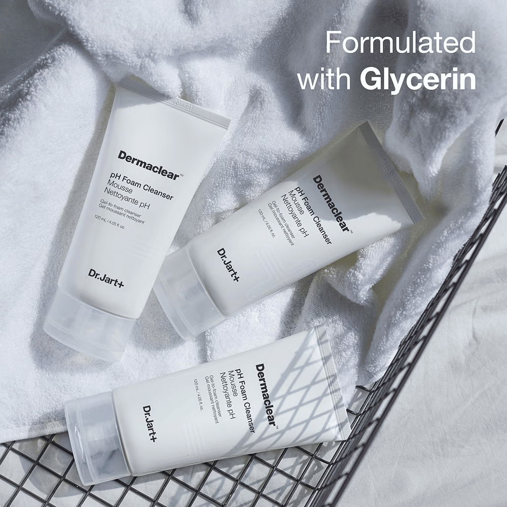 Tre tuber av Dr.Jart+ Dermaclear pH Foam Cleanser ligger på et hvitt frottéhåndkle i en metallkurv, med teksten "Formulated with Glycerin" i øvre hjørnet.