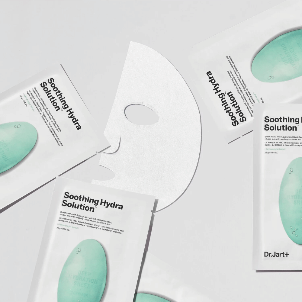 Flere pakker med "Soothing Hydra Solution" ansiktsmasker fra Dr.Jart+ er spredt ut på en lys overflate. En utbrettet maske er plassert i midten, som viser dens hvite og hullstrukturerte utseende.