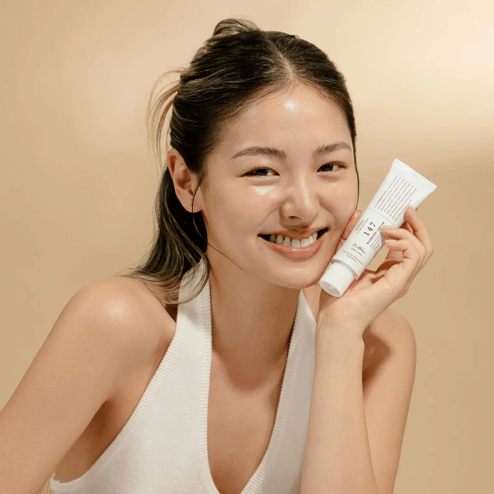 Modell med glødende hud som smiler og holder i Dr. Althea 147 Barrier Cream – en koreansk ansiktskrem for hudbarriere og fuktighet.