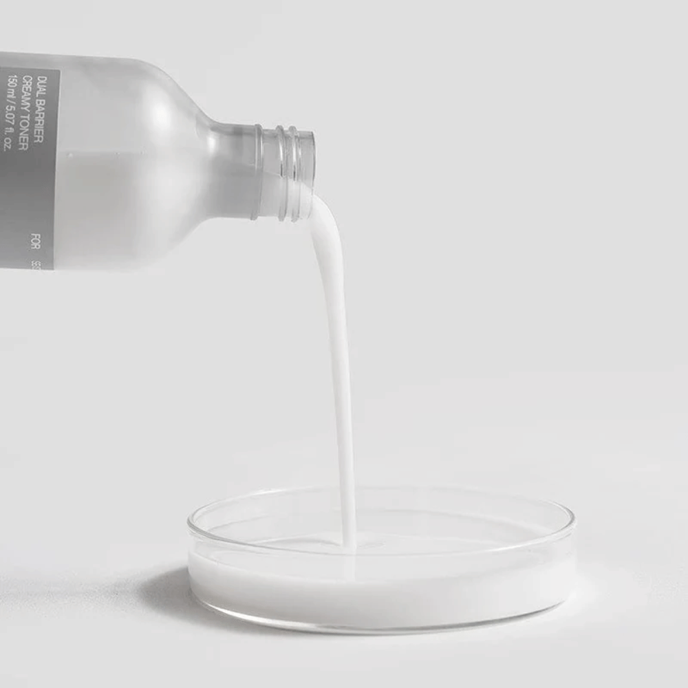Celimax Dual Barrier Creamy Toner helles forsiktig fra flasken ned i en rund laboratoriebrønn, med sin melkete og næringsrike formel tydelig i fokus.