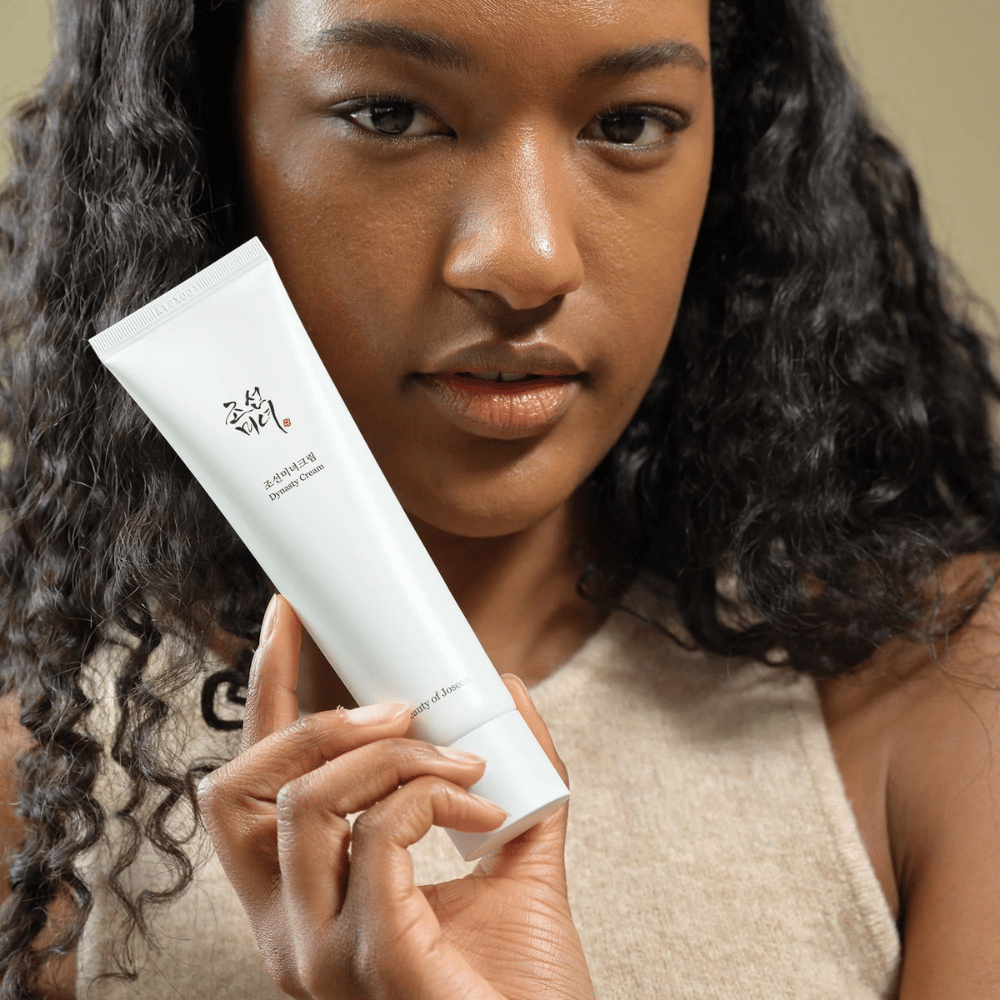 En kvinne med krøllete hår holder en tube "Beauty of Joseon Dynasty Cream" nær ansiktet sitt, noe som fremhever produktet. Kremen, presentert i en stilig hvit tube, er designet for å nære og forynge huden, noe som understreker dens attraktivitet for ulike hudtyper.
