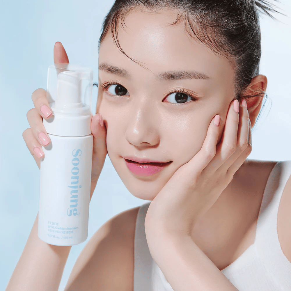 En ung kvinne med klar hud holder en hvit pumpeflaske med teksten "Soon Jung pH 6.5 Whip Cleanser" fra ETUDE. Hun smiler mot kameraet med lett sminke og en frisk utstråling mot en lys blå bakgrunn.