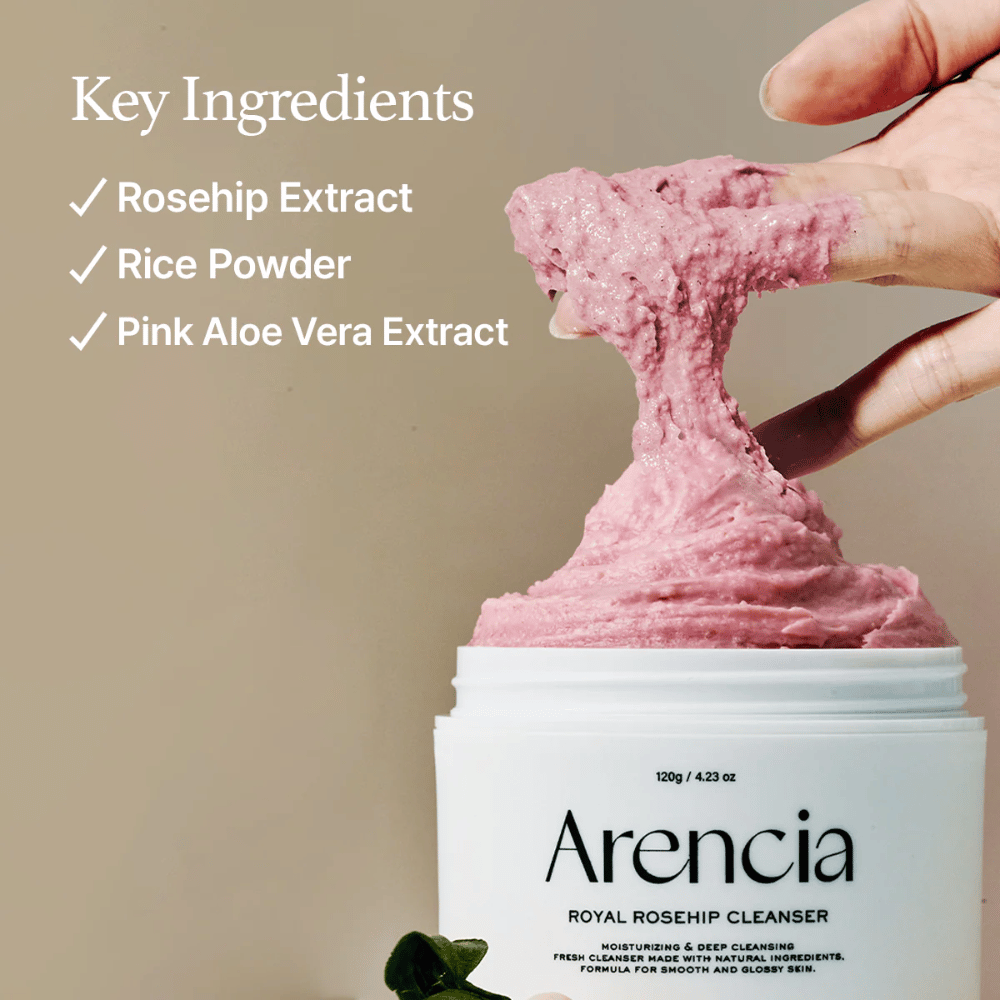 En hånd løfter opp den rosa, fluffy rengjøringsteksturen fra krukken Arencia Royal Rosehip Cleanser, med ingrediensliste til venstre: nypeekstrakt, rispulver og rosa aloe vera-ekstrakt.