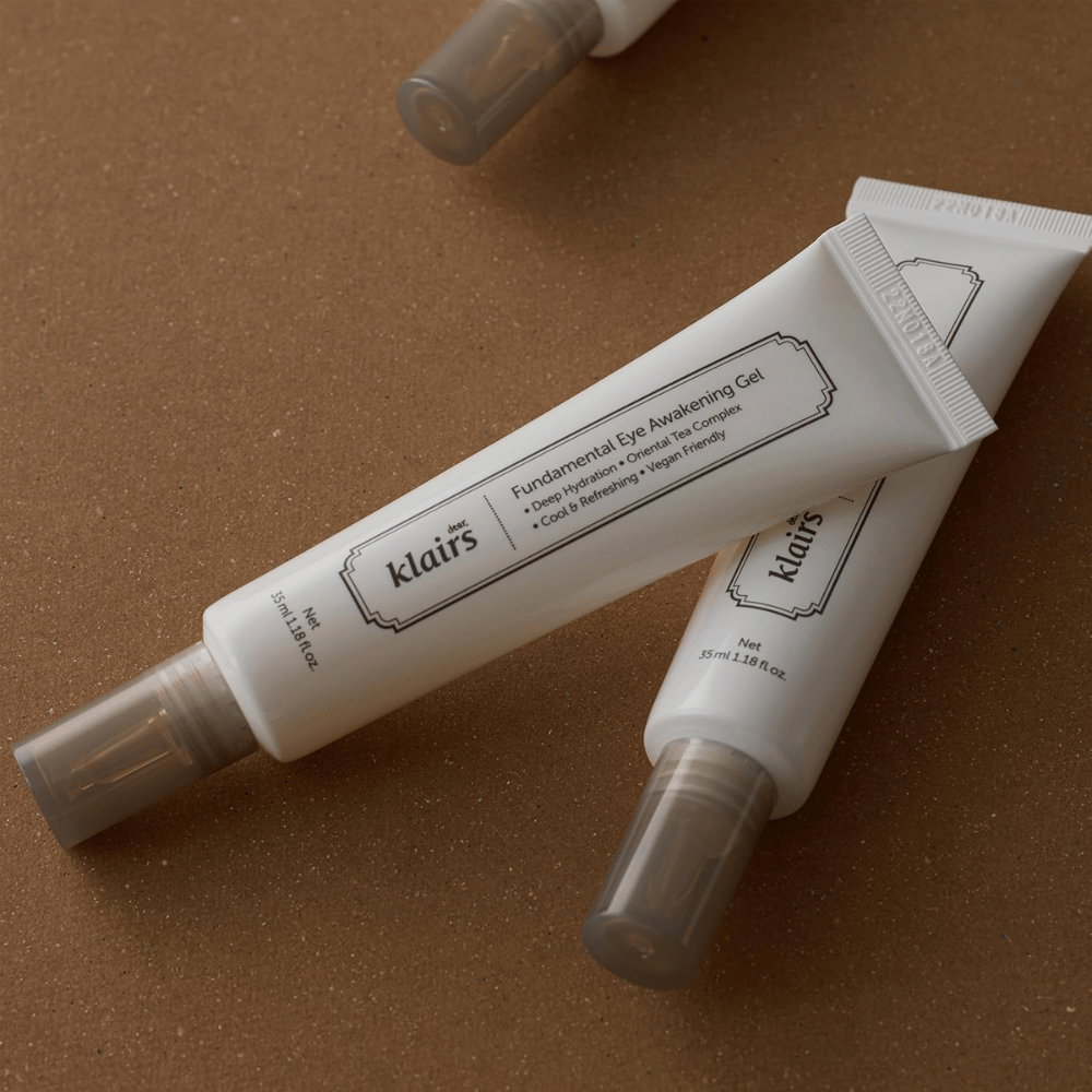 Livsstilsbilde med to tuber av Klairs Fundamental Eye Awakening Gel plassert mot en varm brun bakgrunn. Komposisjonen fremhever produktets elegante design og premiumfølelse samtidig som fokuset ligger på øyepleie og fuktighet.