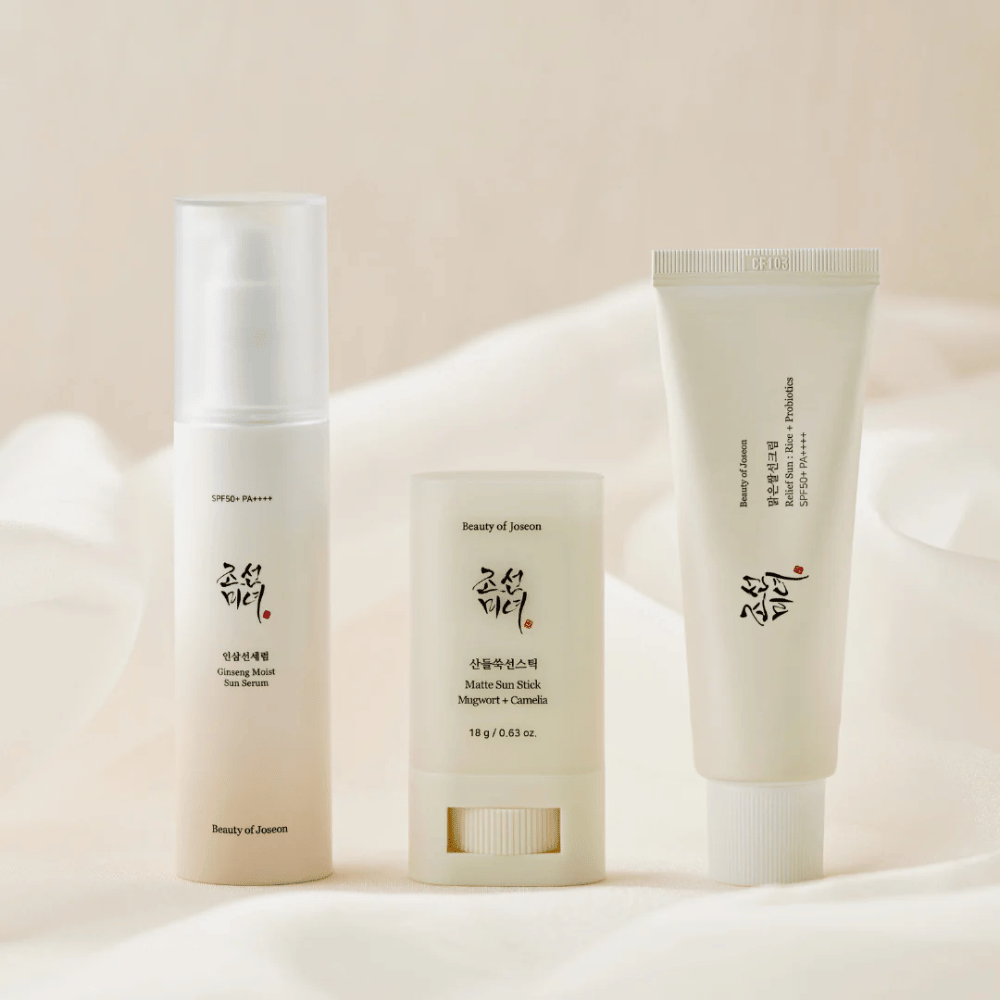 Tre solbeskyttelsesprodukter fra Beauty of Joseon – Ginseng Moist Sun Serum, Matte Sun Stick og Relief Sun Rice + Probiotics – står på rekke på et kremfarget stoff med mykt lys.