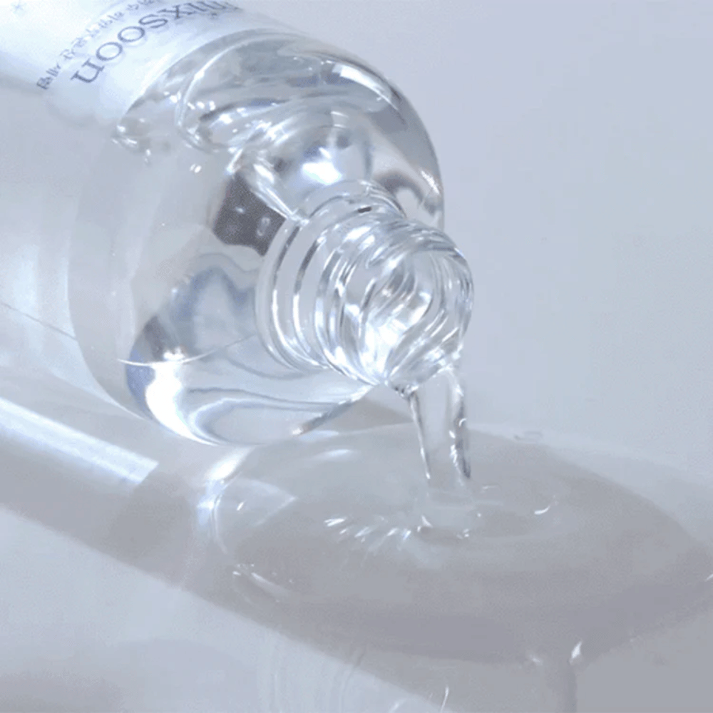Nærbilde av gjennomsiktig serum som helles fra en flaske Mixsoon Glacier Water Hyaluronic Acid Serum på en blank overflate.