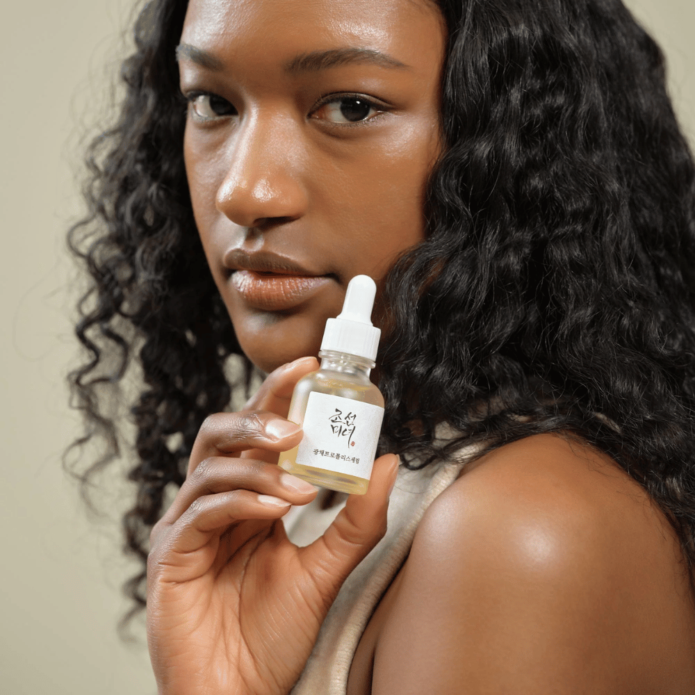 Kvinne med krøllete hår holder opp en flaske Beauty of Joseon Glow Serum med pipette, en fuktighetsgivende og glødgivende hudpleieprodukt.