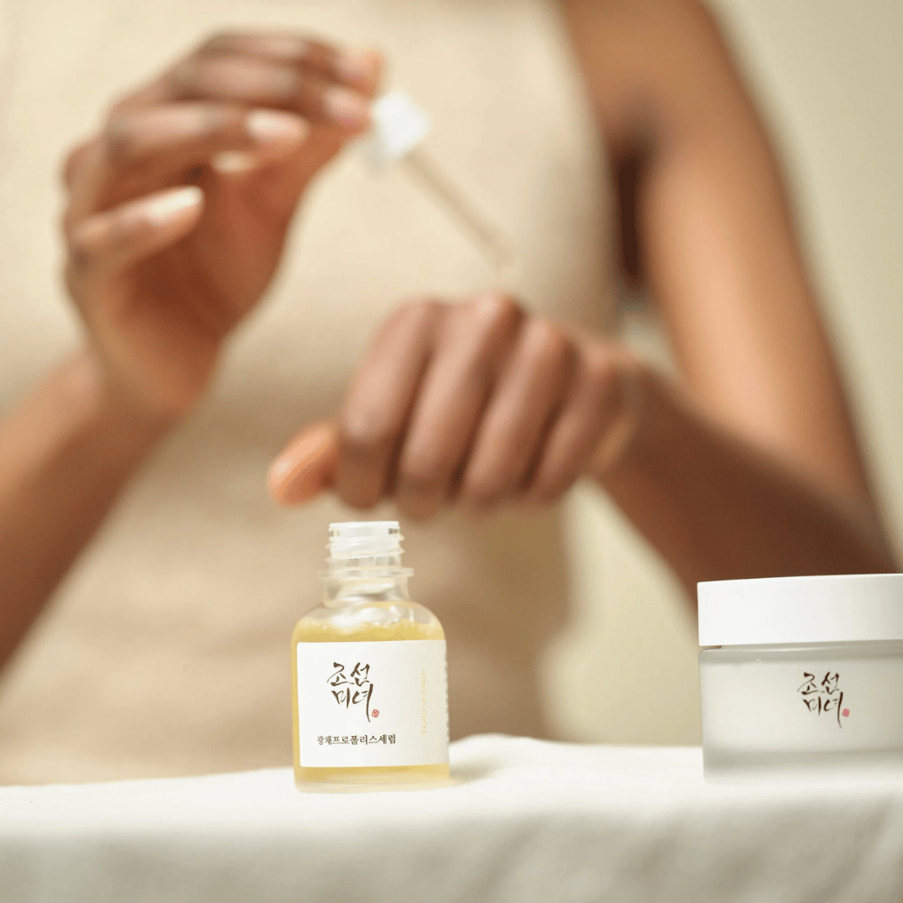 Person som påfører Beauty of Joseon Glow Serum med pipette fra glassflaske, med en krukke av samme serie ved siden av på bordet.