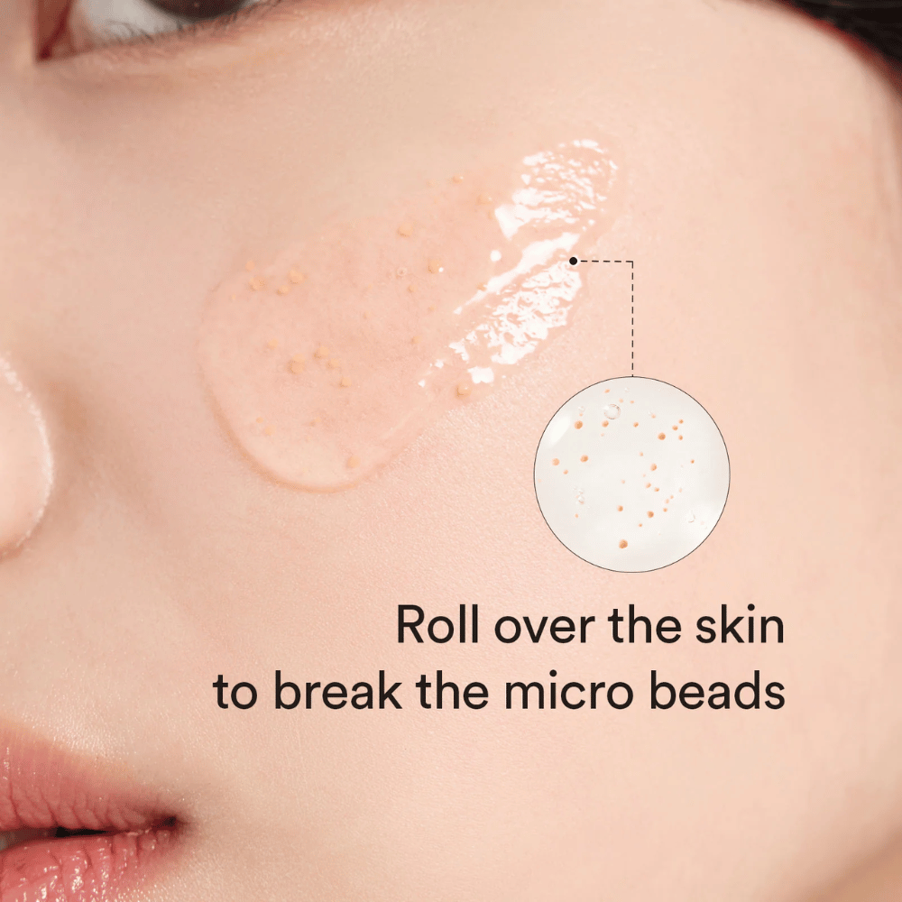 Nærbilde av et kinn med et serum som inneholder små mikropartikler påført huden. En sirkelzoom viser partiklene tydeligere. Teksten sier "Roll over the skin to break the micro beads".