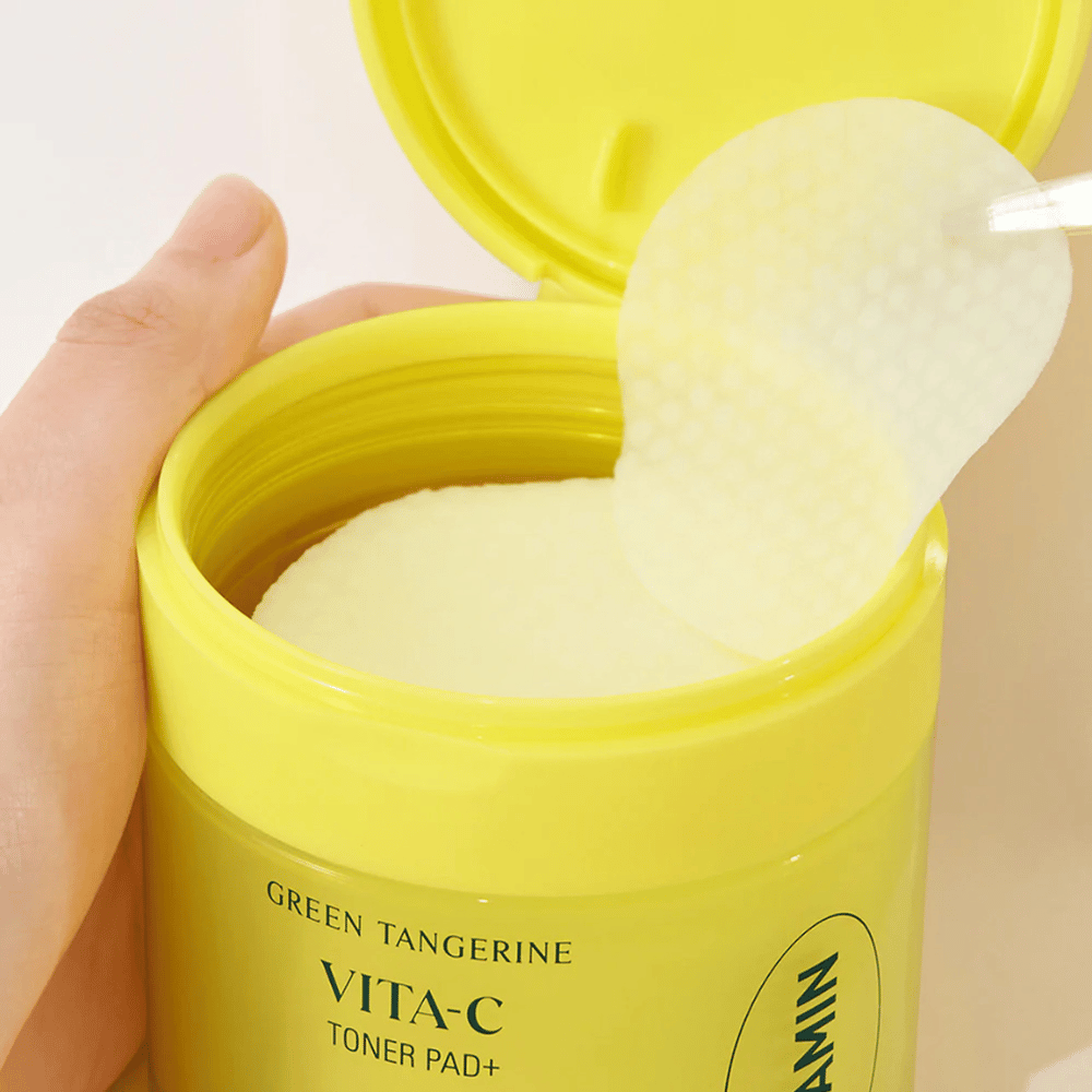 En hånd bruker en plastpinsett for å plukke opp en tonerpad med tekstur fra en lysegul beholder merket "GREEN TANGERINE VITA-C TONER PAD+" av merket "gooda