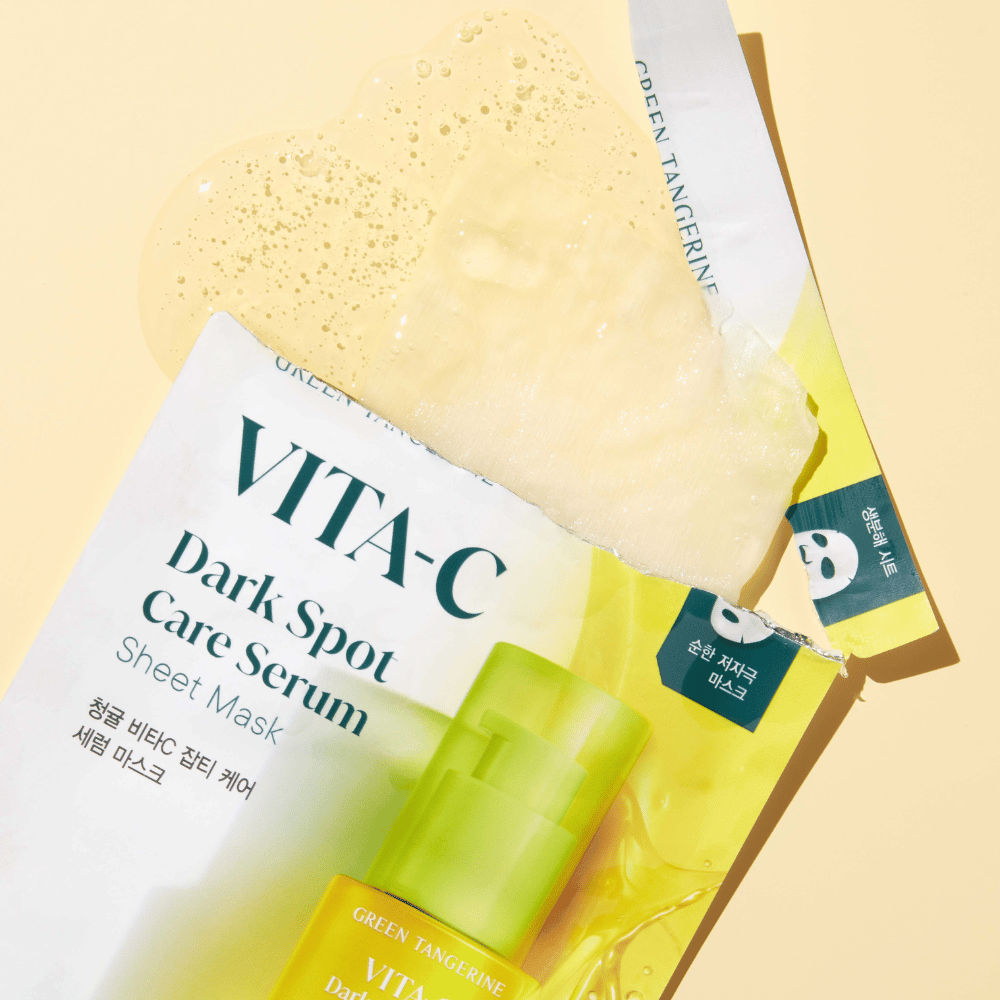 Åpnet emballasje av VITA-C Dark Spot Care Serum Sheet mask med en ansiktsmaske på toppen og en grønn serumflaske, alle mot en gul bakgrunn.
