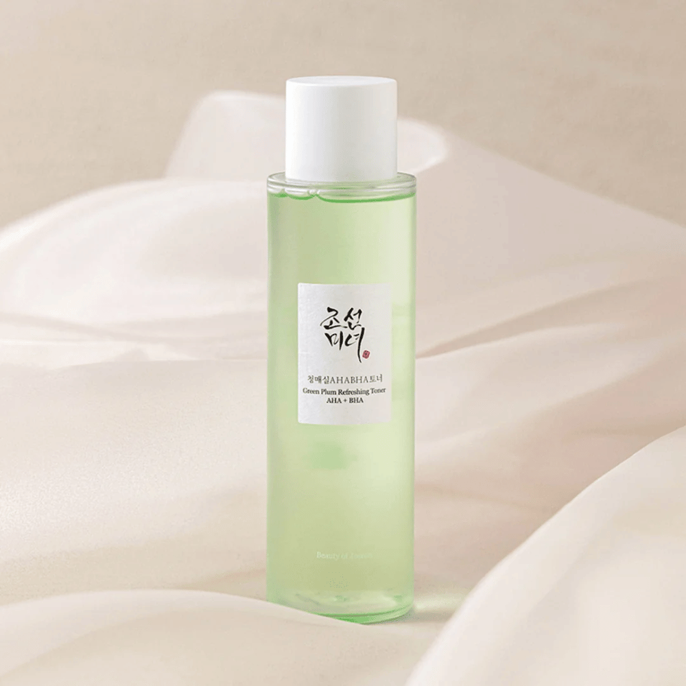 En flaske med "Green Plum Refreshing Toner" AHA + BHA står på en myk, lys bakgrunn. Produktet har en grønn væske som reflekterer friskhet og naturlighet. Etiketten er hvit med svarte og røde innslag, noe som gir en elegant og ren look.