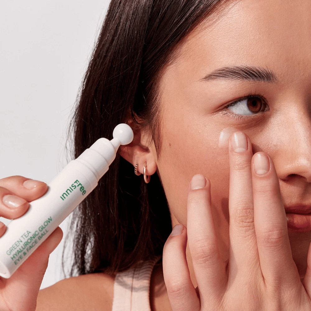 En person påfører Innisfree Green Tea Hyaluronic Glow Eye & Face Ball på huden under øynene. Produktet har en hvit, rund applikasjonsdel og er i en hvit flaske med grønn tekst. Brukerens ansikt er delvis synlig og fokuserer på påføringen av produktet.