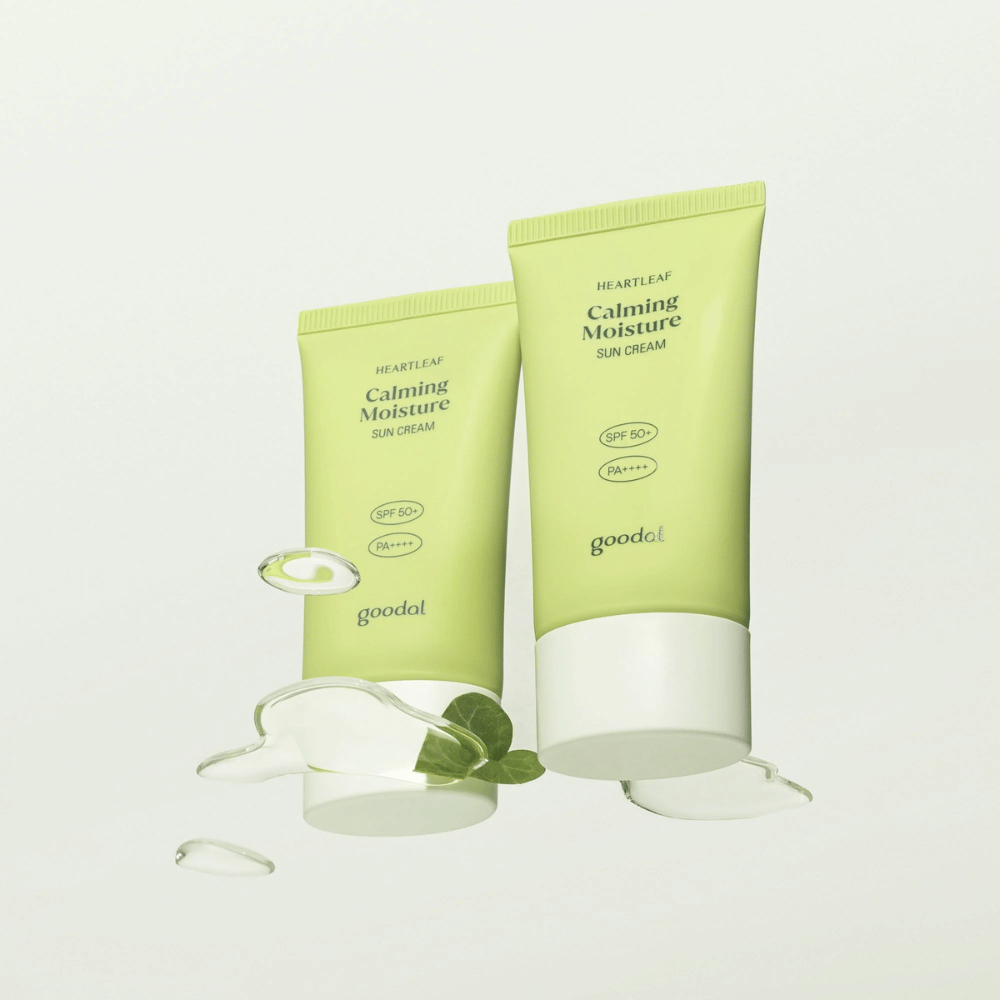 To tuber av Goodal Heartleaf Calming Moisture Sun Cream SPF50+ PA++++ omgitt av planteblader og serumdråper mot lys bakgrunn.