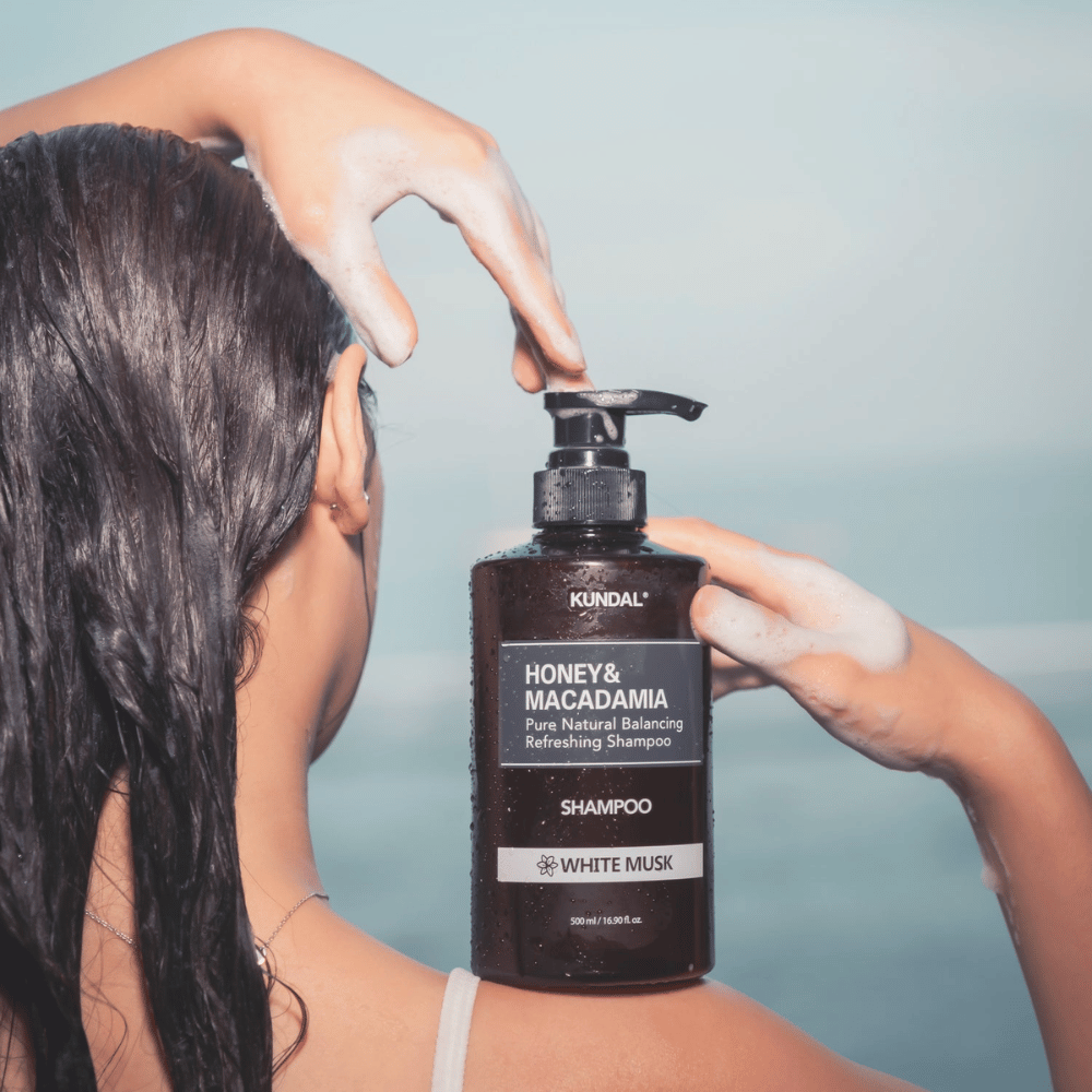 Kvinne med vått hår påfører skum og holder KUNDAL Honey & Macadamia Nature Shampoo White Musk mot havbakgrunn.