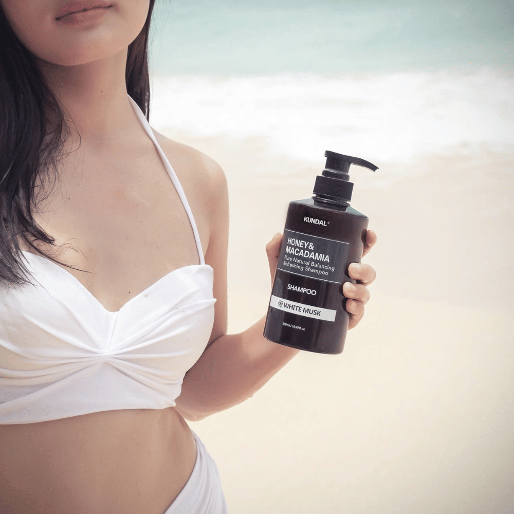 Kvinne i hvit bikini holder KUNDAL Honey & Macadamia Nature Shampoo White Musk på en strand med havet i bakgrunnen.