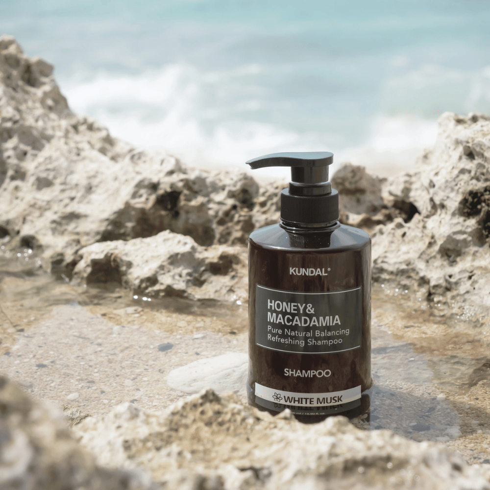 KUNDAL Honey & Macadamia Nature Shampoo White Musk plassert på klipper ved havet, et naturlig hårpleieprodukt med frisk duft.