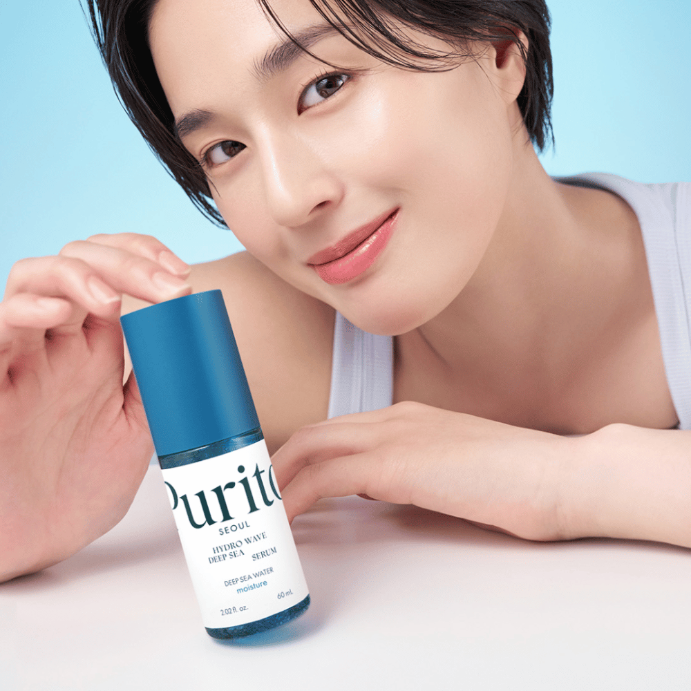 En kvinne med kort hår holder et blått serum fra Purito, Hydro Wave Deep Sea Serum. Hennes hud ser strålende og fuktig ut, noe som fremhever produktet i fokus. Bakgrunnen er lyseblå, noe som gir en frisk og ren følelse. Produktet er fordypet i hudpleie.