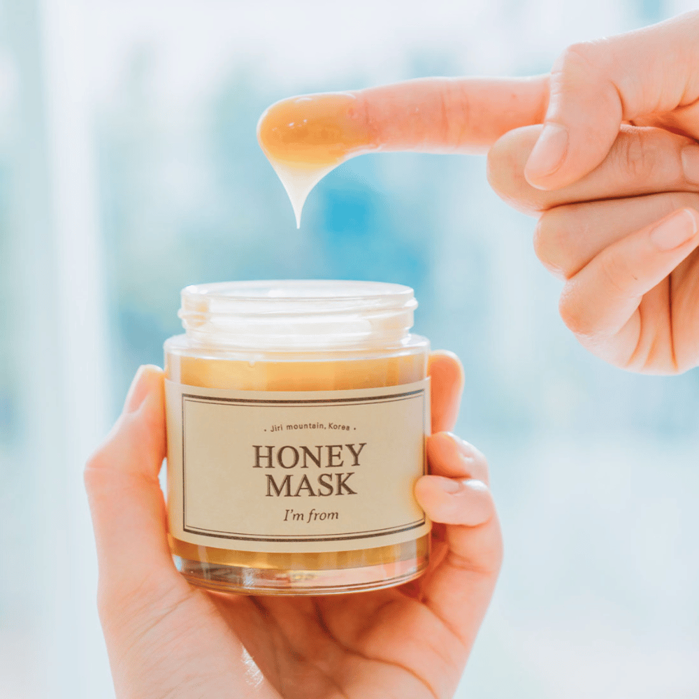 En hånd holder åpen boks av "I'm from Honey Mask", og en finger dyppes i den gyldne masken mot en lys bakgrunn.