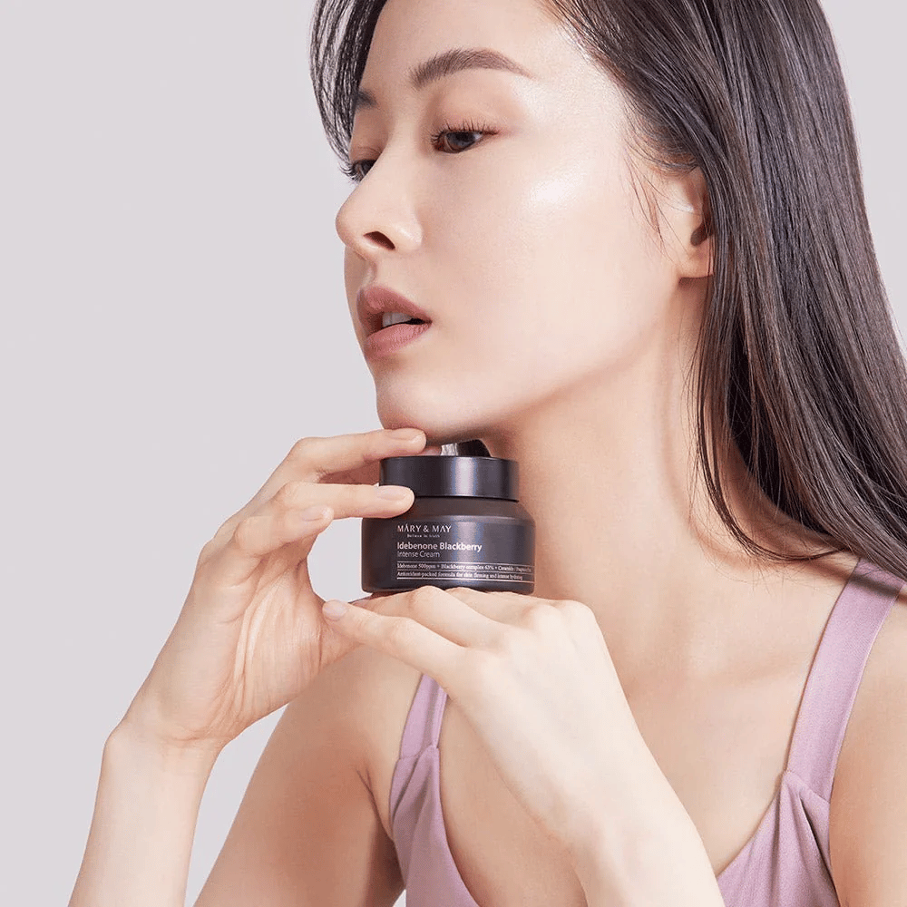 En kvinne holder en boks med MARY & MAY Idebenone Blackberry Intense Cream. Hun har en strålende hud og smiler lett, kledd i en ermeløs topp. Produktet er en intensiv krem med idebenon og bjørnebærkompleks som er ment for hudens fasthet og fuktighet.