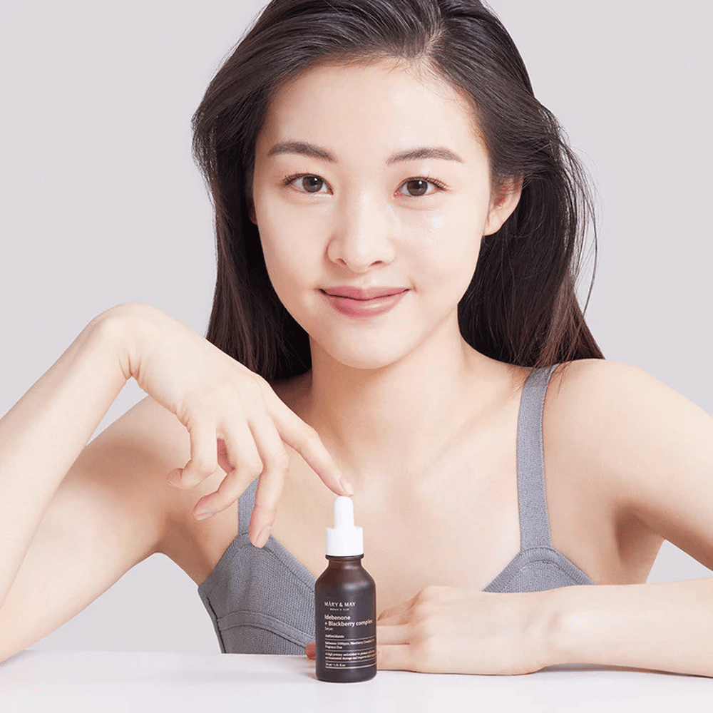 En kvinne med langt, mørkt hår holder i MARY & MAY Idebenone + Blackberry Complex Serum, en mørk glassflaske med pipette. Hun har et bredt smil og peker på flasken med den andre hånden. Kvinnen er kledd i en grå, ermeløs topp og ser ut til å befinne seg i et lyst og enkelt miljø, som fremhever produktets fokus på hudpleie og skjønnhet.