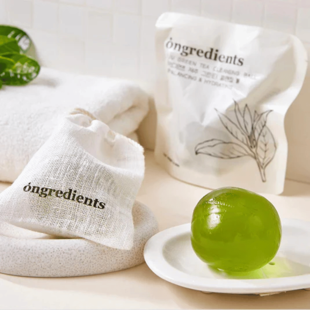 Óngredients Jeju Green Tea Cleansing Ball plassert på fat med emballasje og håndkle – skånsom rengjøring med grønn te fra Jeju.
