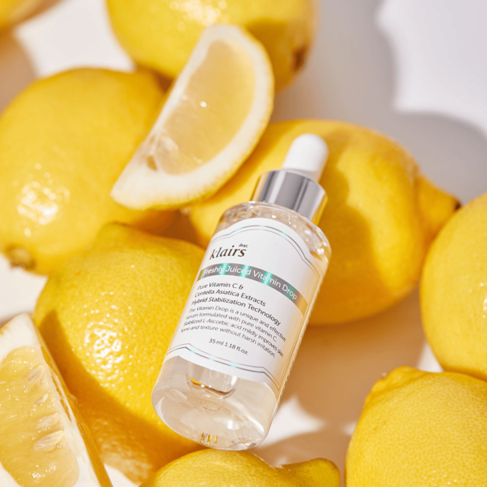 Klairs Freshly Juiced Vitamin Drop serumflaske omgitt av ferske sitronhalvdeler og sitronskiver på en lys overflate, fremhever ingrediensene.
