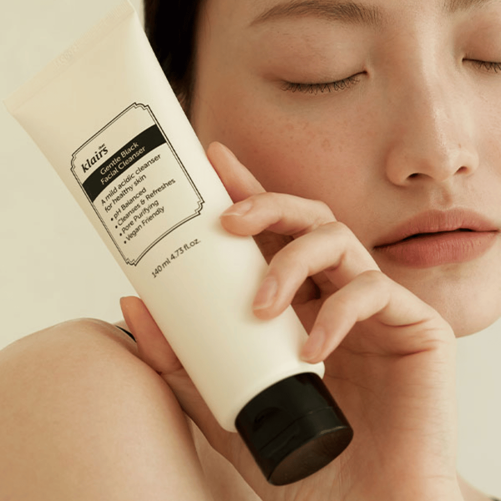 En person holder en tube av Klairs Gentle Black Facial Cleanser mot sitt kinn, lukkede øyne og naturlig hudtone vises i bakgrunnen.