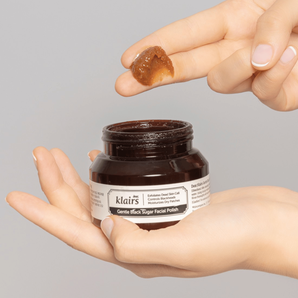 Hender holder en åpen krukke av Klairs Gentle Black Sugar Facial Polish og viser produktet på en finger, mot en grå bakgrunn.