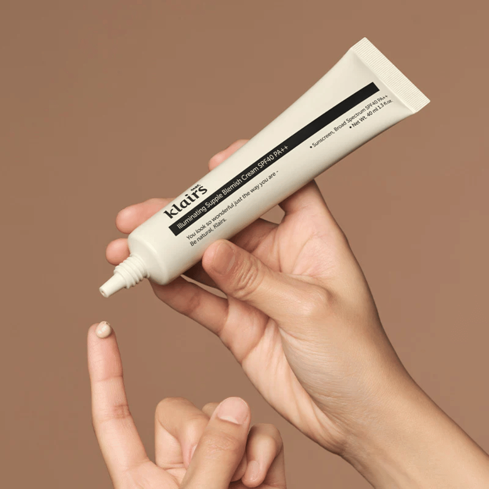 En hånd holder en tube av Klairs Illuminating Supple Blemish Cream SPF40 PA++ og trykker ut en liten mengde krem på en finger, mot en brun bakgrunn.