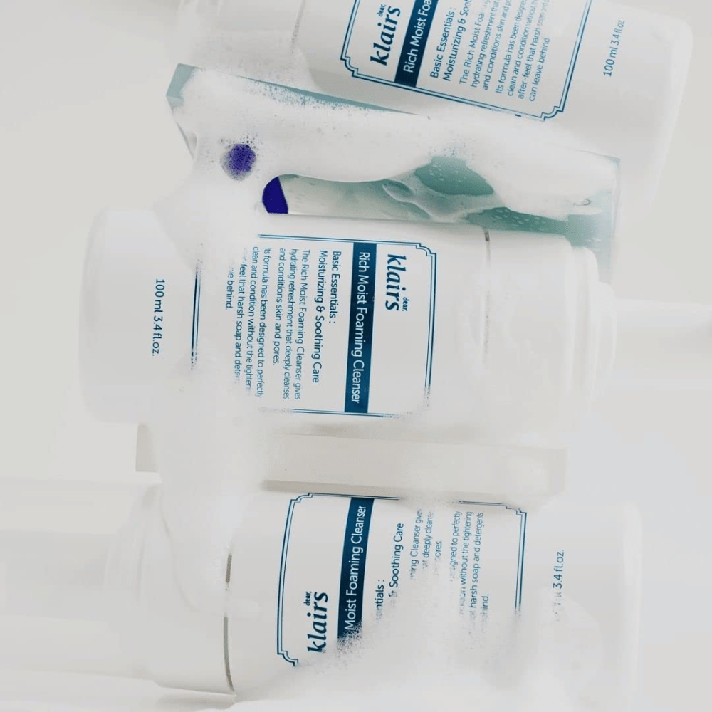 Tre flasker av Klairs Rich Moist Foaming Cleanser dekket med skum og vanndråper, plassert på en hvit overflate.