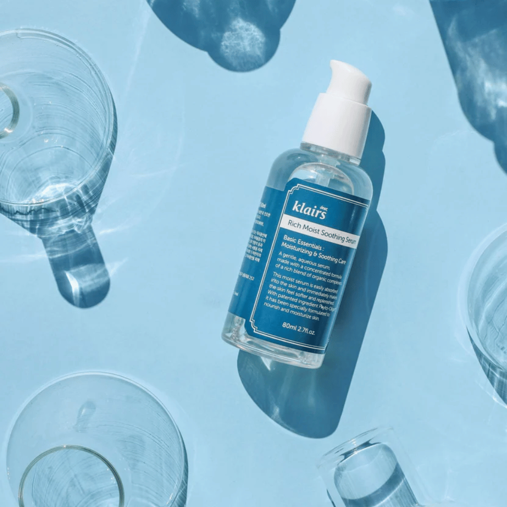 En flaske Klairs Rich Moist Soothing Serum blant vannglass med refleksjoner på en blå bakgrunn.