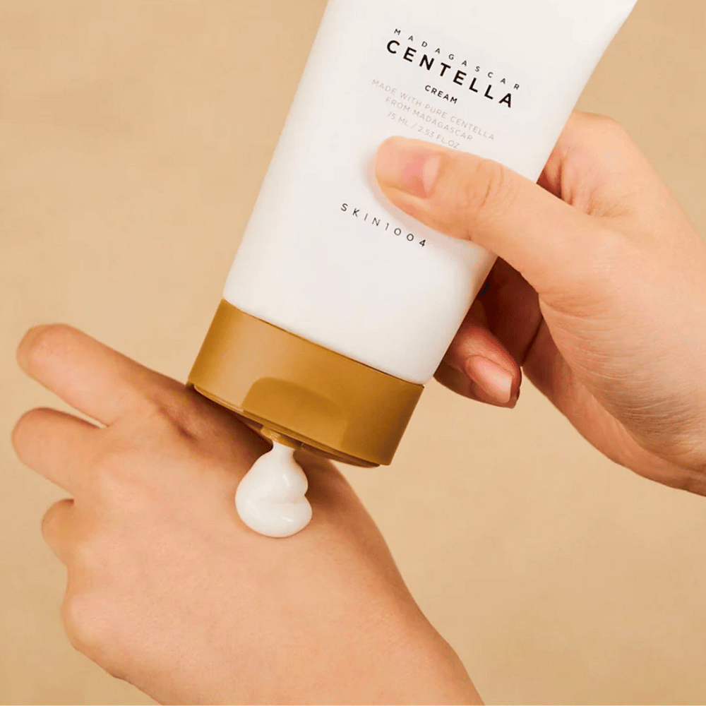 Bildet viser en person som holder i en tube av "Madagascar Centella Cream" fra SKIN1004. Det er en hudkrem som er laget av ren Centella fra Madagaskar, i størrelsen 75 ml / 2.53 fl.oz. Kremen påføres på hånden og er ment å gi næring og fuktighet til huden.