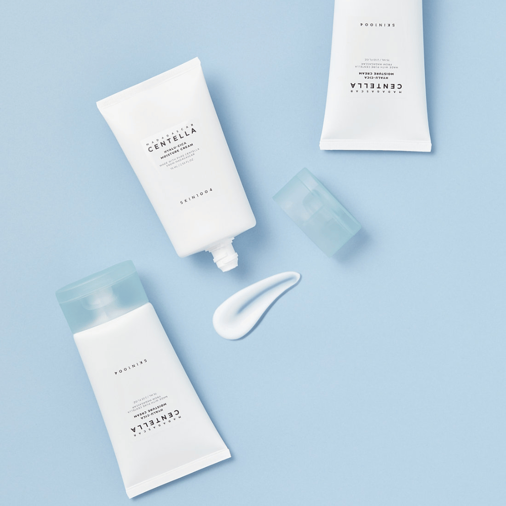 To hvite tuber med SKIN1004 Centella hudpleieprodukter, en klatt av produkt og en klar blå kapsyl, på en lys blå bakgrunn.